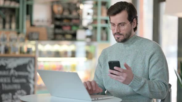 Young Man with Laptop Checking Smartphone in Cafe