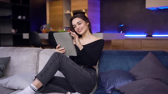 Modern Young Brunette Woman in Black Top and Jeans Sitting on the Home Couch and Using Tablet PC