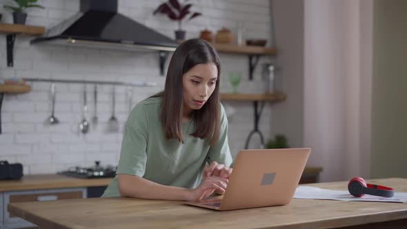 Beautiful Young Asian Woman Holding Head in Hands Messaging Online on Laptop in Home Office
