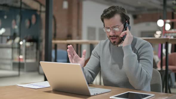 Angry Young Man Talking on Smartphone While Using Laptop in Office