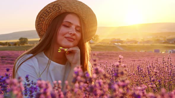 Beautiful Young Girl in a Straw Hat and White Dress Sitting in a Lavender Field on Sunset