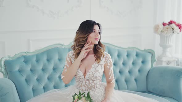 Gorgeous young woman on the couch with her hand on neck. Luxurious bride in a wedding dress