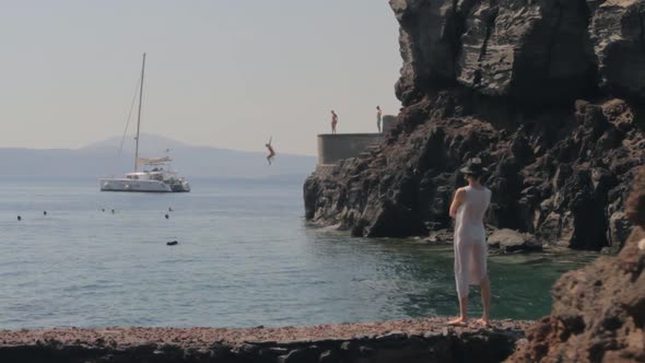 Woman watching a dive from a cliff in a rocky beach. A catamaran hovers nearby, between the dark cli