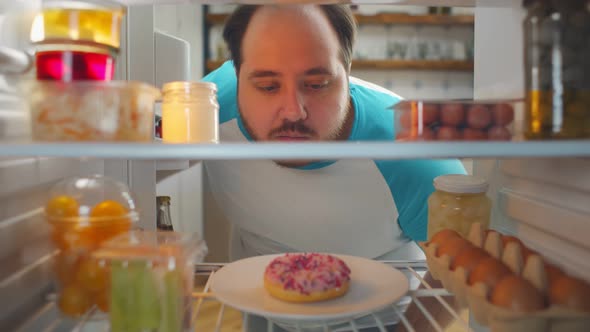 View From Fridge of Stout Young Man Looking at Donut but Choosing Fresh Vegetables in Container.