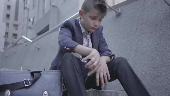 Sad Well-dressed Boy Sitting on the Stairs on the Street, Old Purse Near Him