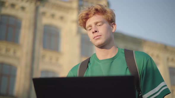 Concentrated Redhead Man Sitting in Sunlight Outdoors and Using Laptop. Portrait of Positive Focused