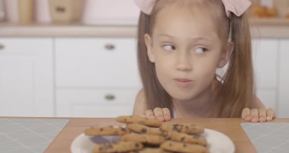 Cheerful Little Girl Peeking Out of Table and Taking Sweet Tasty Cookie From Plate