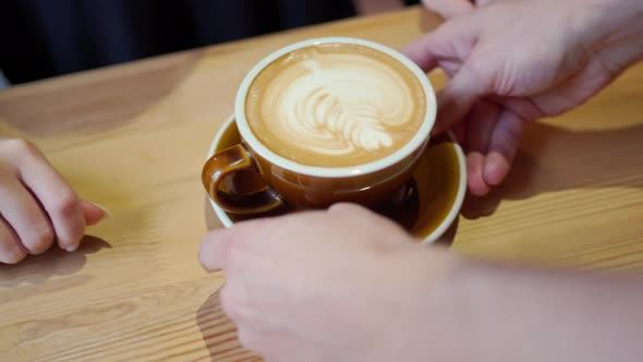 Barista's Hands Put a Brown Cappuccino Mug with Beautiful Latte Art on the Table Where Female Hands