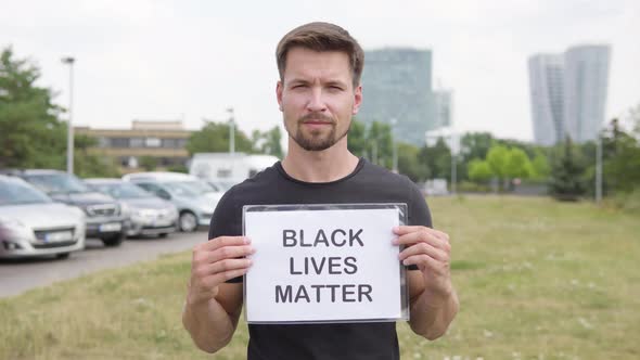 A Young Handsome Caucasian Man Shows a Black Lives Matter Sign To the Camera By a Parking Lot