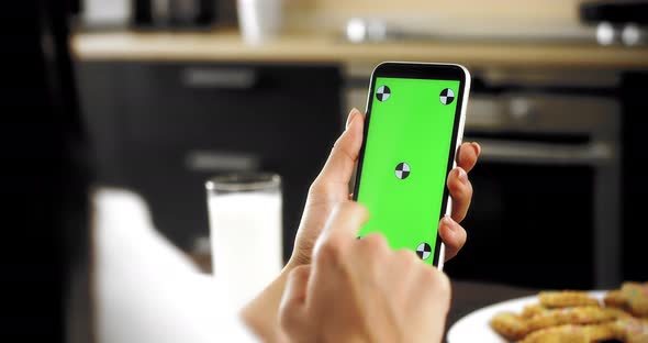 Woman Holds a Smartphone with a Green Screen in Her Hands Sitting in the Kitchen