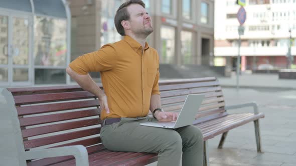 Young Man with Back Pain Using Laptop while Sitting Outdoor on Bench