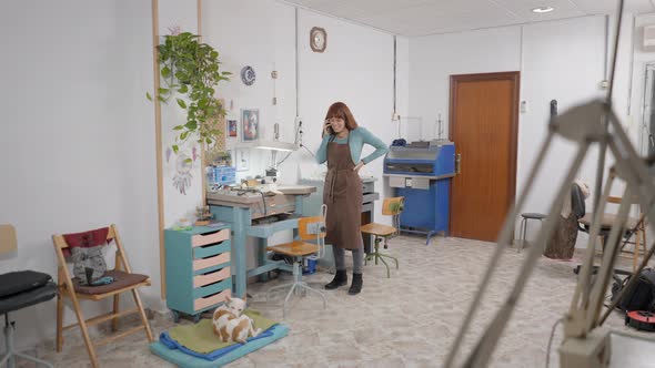 Young Female Entrepreneur Talking On Her Smartphone Inside The Shop Pet Dog Grooming Himself On Dog