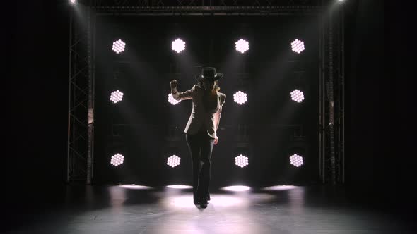 An Attractive Woman in a Pantsuit and Hat Walks in a Dark Studio Against the Backdrop of Neon Lights