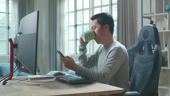 Asian Man Using Mobile Phone And Drinking Coffee While Using Computer For Working At Home