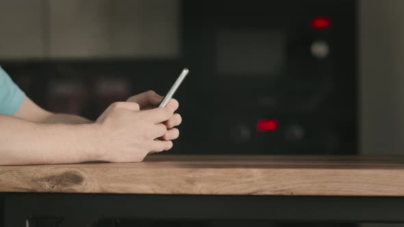 A Man with a Mobile Phone in His Hands Stands Near a Wooden Table Uses a Mobile Application