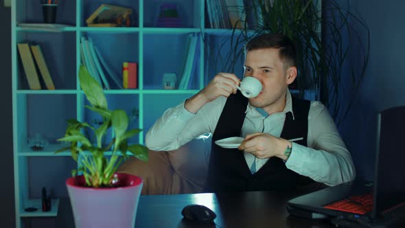 Young Man Drinks Coffee Sitting in Armchair Before Computer in Office