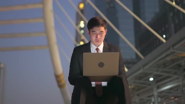 Asian business man working on computer notebook sitting outdoor at night in city for urgent job.