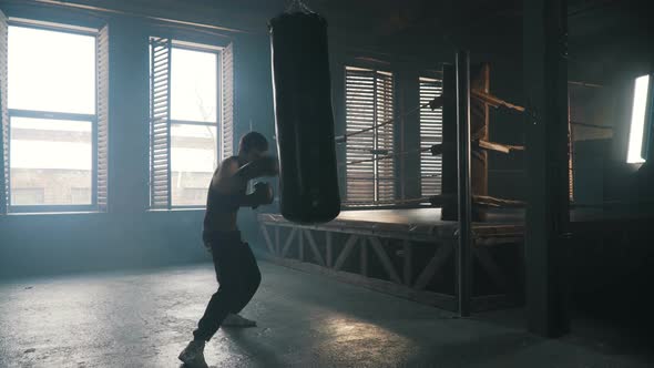 Young Man Punch The Bag Hard Near Boxing Ring In Industrial Interior Silhouette