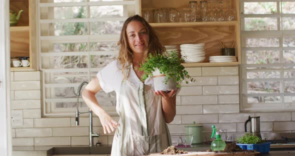 Portrait of smiling caucasian woman holding potted plant, standing in sunny cottage kitchen