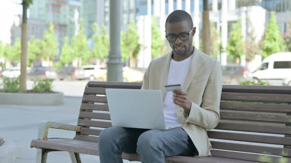 Excited African Man Shopping Online on Laptop Sitting Outdoor on Bench