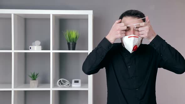 Doctor in a Black Shirt Puts a Protective Mask on His Face in a Cozy Office and Looks at the Camera