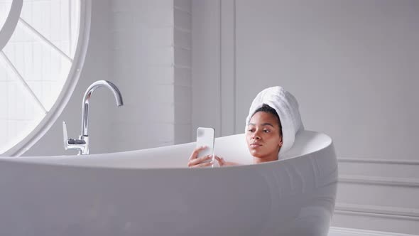 Black Woman Taking a Bath Looking at Phone with Her Hair Wrapped in a Towel