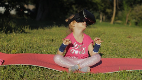 Child in VR Headset Helmet Sitting in Lotus Position on Mat and Performing Yoga Meditation in Park