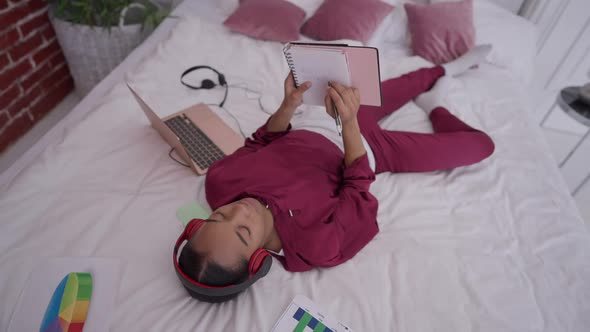 Young Woman Writing Remote Working in Sketchpad Lying on Bed in Bedroom