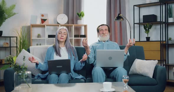 Mature Couple Sitting on Soft Couch at Home with Computers and Meditating with Closed Eyes