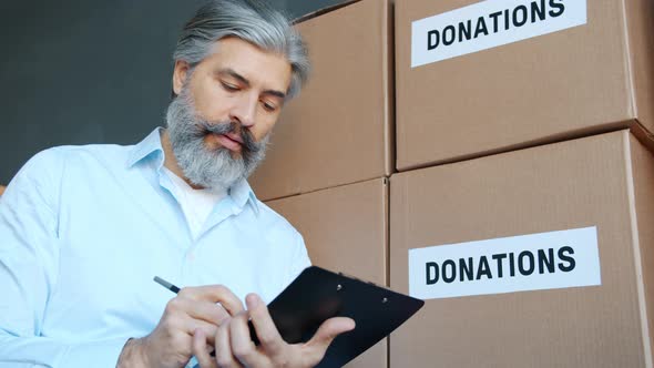 Mature Man Charity Company Manager Doing Paperwork in Storage Room with Donation Boxes