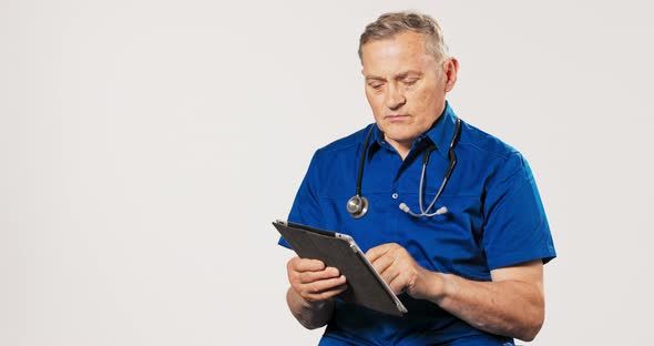 Professional Mature Doctor Dressed in Blue Uniform Gown and Stethoscope Around His Neck Clicks Away