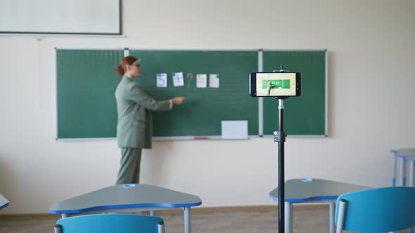 Online Math Lesson Teacher in Glasses with Cards Near Blackboard Records a Video Using Mobile Phone