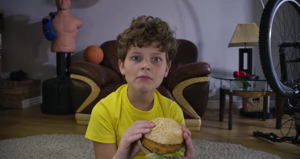 Face of Caucasian Curly-haired Boy Chewing Hamburger and Watching TV. Cute Teenager with Fast Food