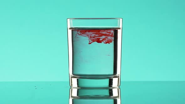 Drops of Red Ink Dripping Into in Glass with Clear Water Going Round in Circles on Turquoise