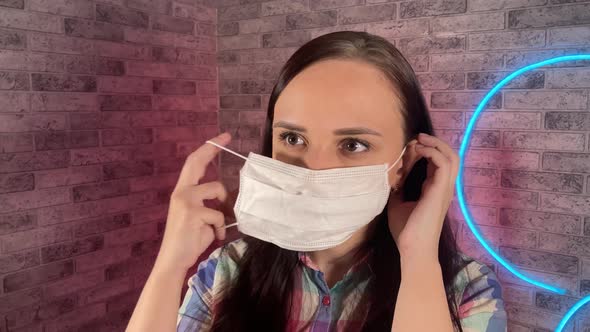 Portrait of Attractive Woman Putting Medical Mask on Face Against Illuminated Wall at Home. Close Up