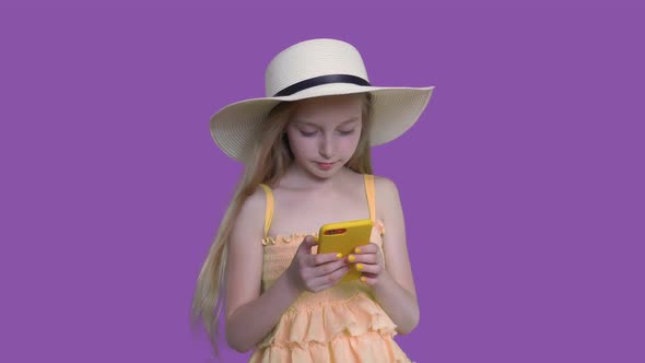 Blonde Child Girl in Summer Hat Is Browsing Smartphone on Purple Wall Background