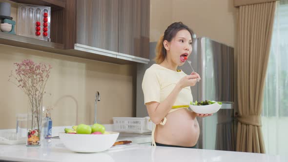 Asian beautiful pregnant woman eating vegetables salad healthy food in kitchen at home.