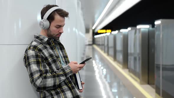 Young Man Waits for a Train in the Subway Uses a Smartphone and Listens to Music with Wireless