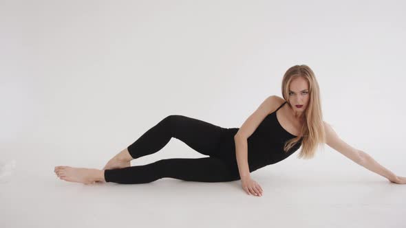 Sensual Young Blonde Girl in Black Tight Sportswear Doing Leg Stretch Lying on the Floor in a White