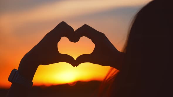 Silhouette Woman Making a Heart Shape with Her Hands Standing in a Field at Sunset