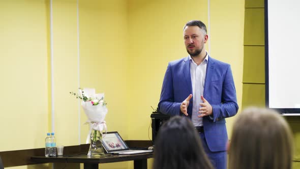 Successful leader is conducting a lecture while standing near table with flowers and a laptop