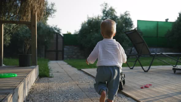 Little Boy Walks on Road Towards Scooter Standing in Yard