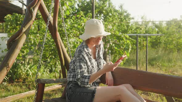 A woman looks into a smartphone, swinging on a swing in the garden.