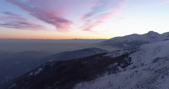 Aerial View of Snowy Mountains with Pink and Orange Sunrise. Fog Over Rolling Below Majestic Snowy