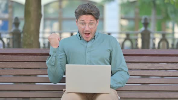 Middle Aged Man Celebrating while using Laptop, Outdoor on Bench