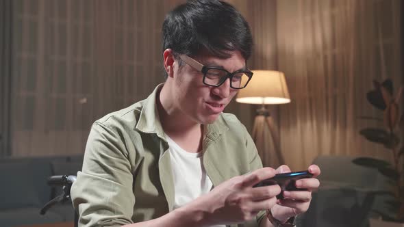 Asian Man Sitting In A Wheelchair While Use Mobile Phone Playing Video Game At Home