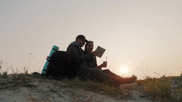 A Young Couple of Tourists Enjoy a Tablet Sitting on Top of a Mountain During Sunset.