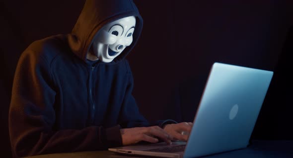 Hacker with masked face uses laptop to hack data.