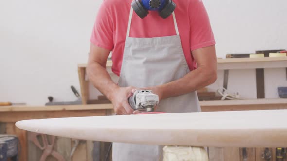 Caucasian male surfboard maker wearing a face mask and preparing to polishing a surfboard