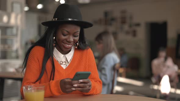 Cheerful African Woman Having Good News on Smartphone While Sitting at Cafe Table